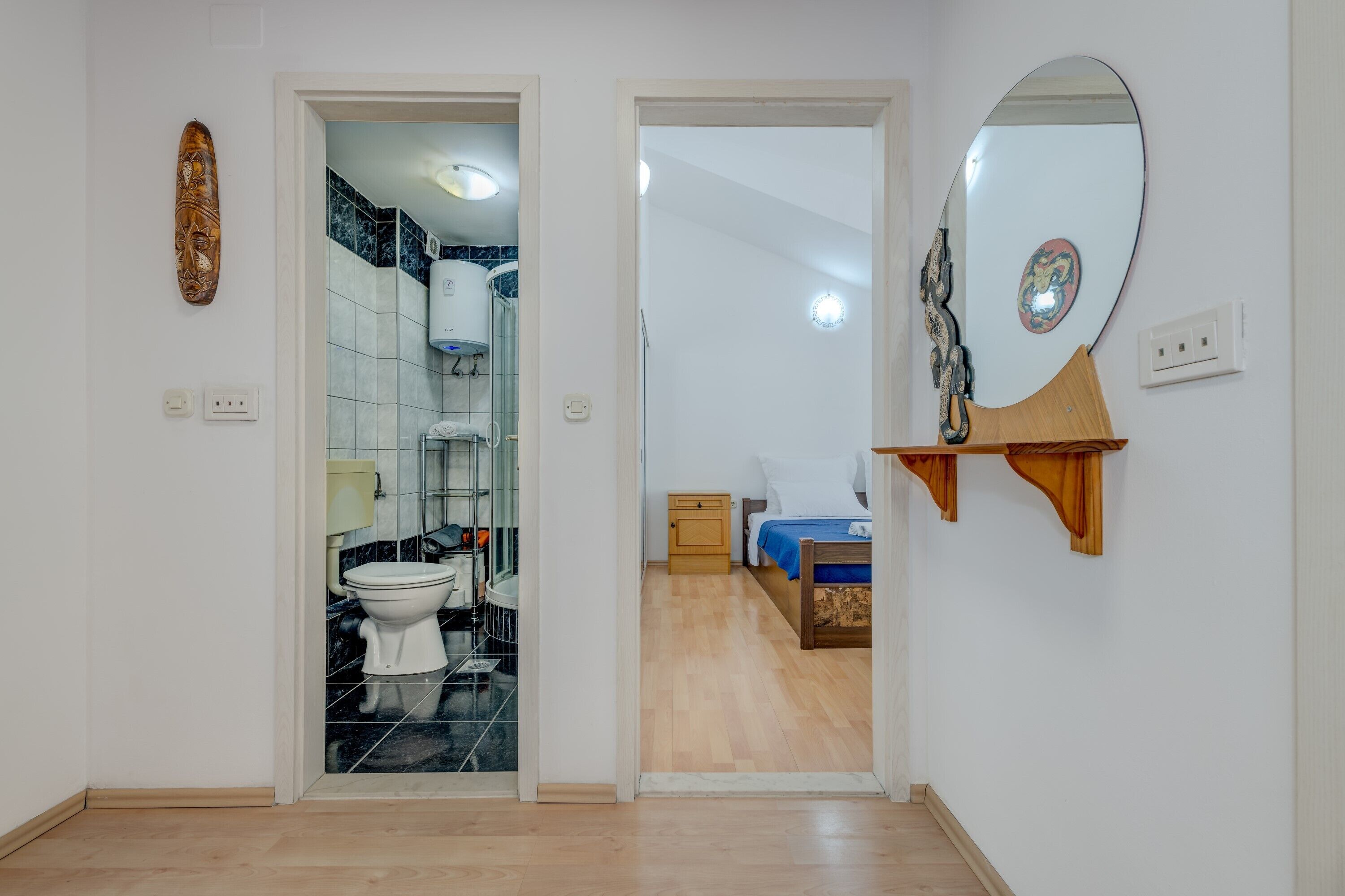 A hallway leading to a bathroom with a toilet and a shower, and a bedroom with a bed and a nightstand.
