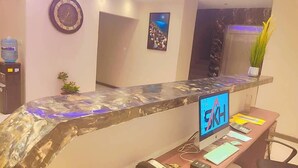 Un mostrador de recepción con un monitor de computadora que muestra 'SKH', un reloj, una obra de arte enmarcada y un dispensador de agua.