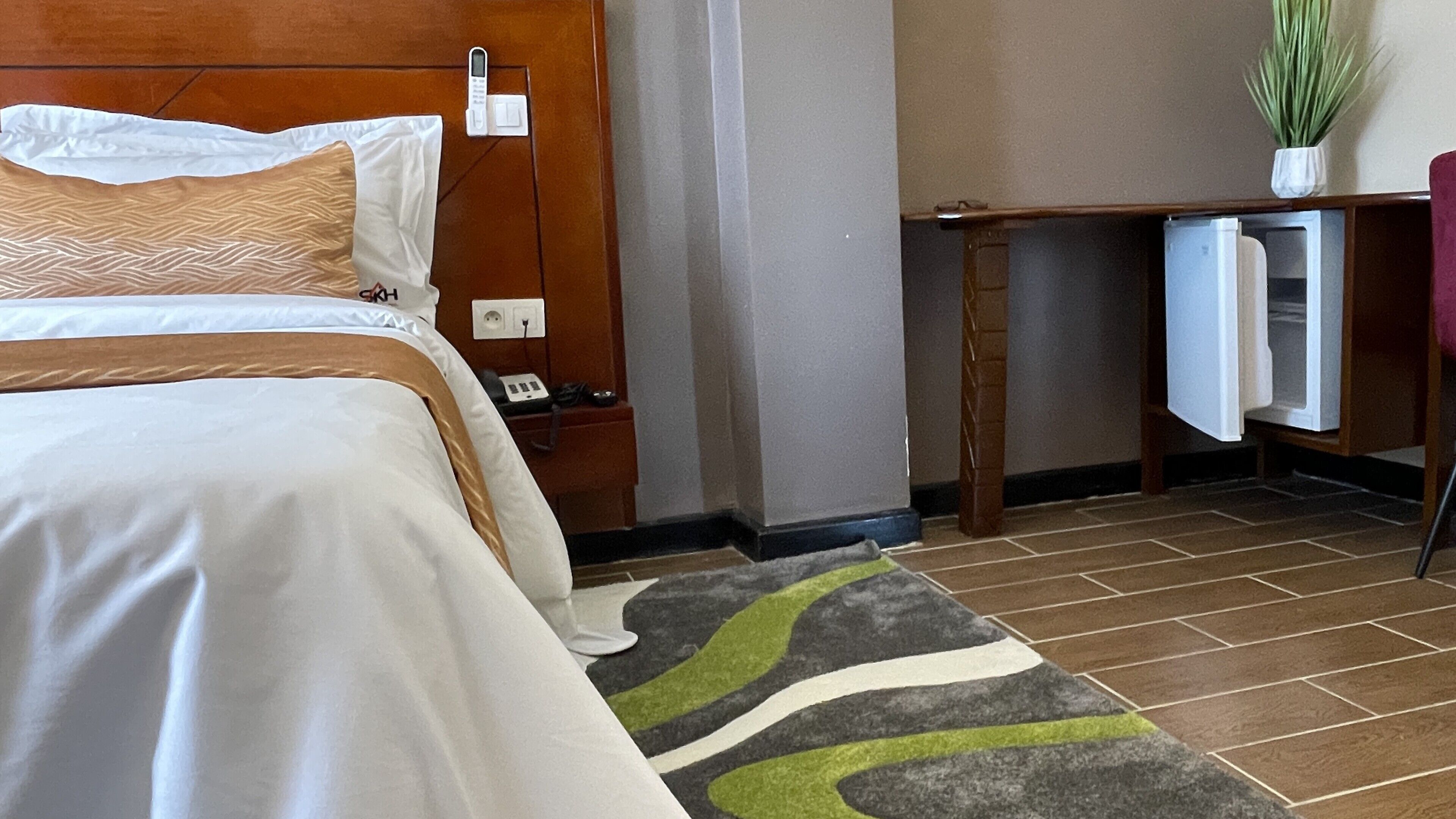 A hotel room with a bed, a desk, a chair, a plant, and a refrigerator.