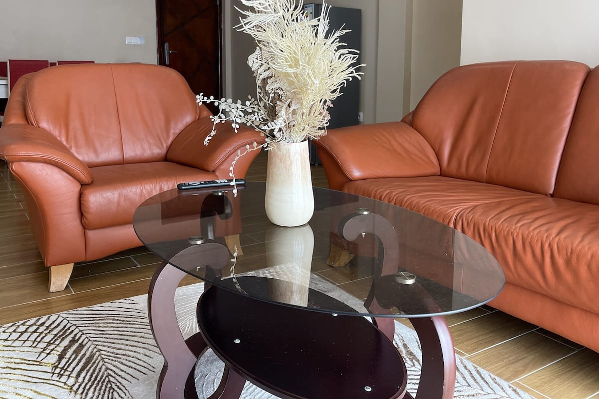 Un salon avec un canapé en cuir marron, une table basse en verre et un vase contenant des plantes séchées.