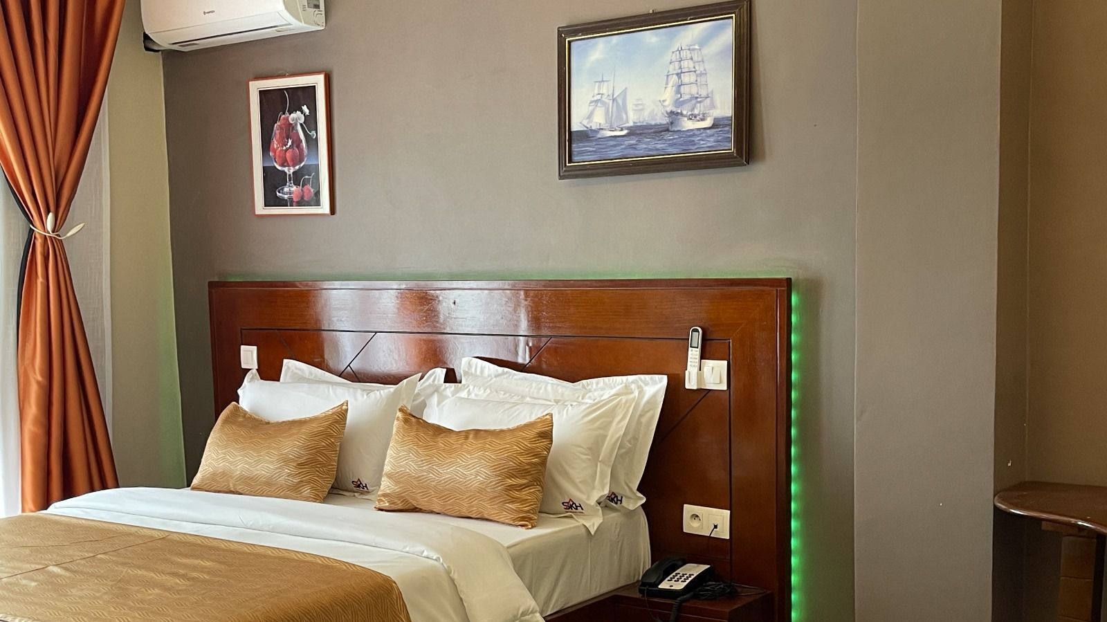 Hotelkamer met houten hoofdeinde, een bed met witte beddengoed en gestreepte kussens, een nachtkastje met telefoon, en twee ingelijste foto’s aan de muur.