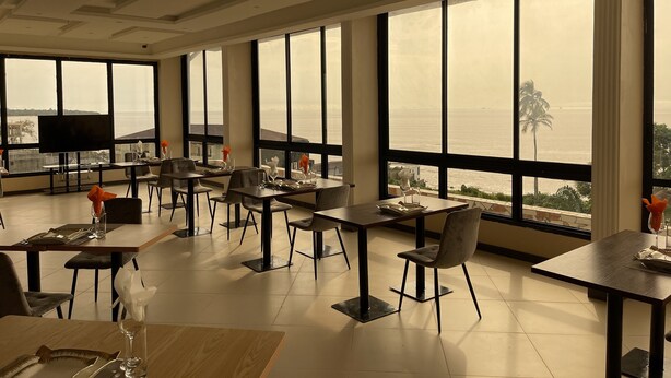 Un comedor con mesas y sillas, ventanas amplias con vista al mar y un televisor en la pared.