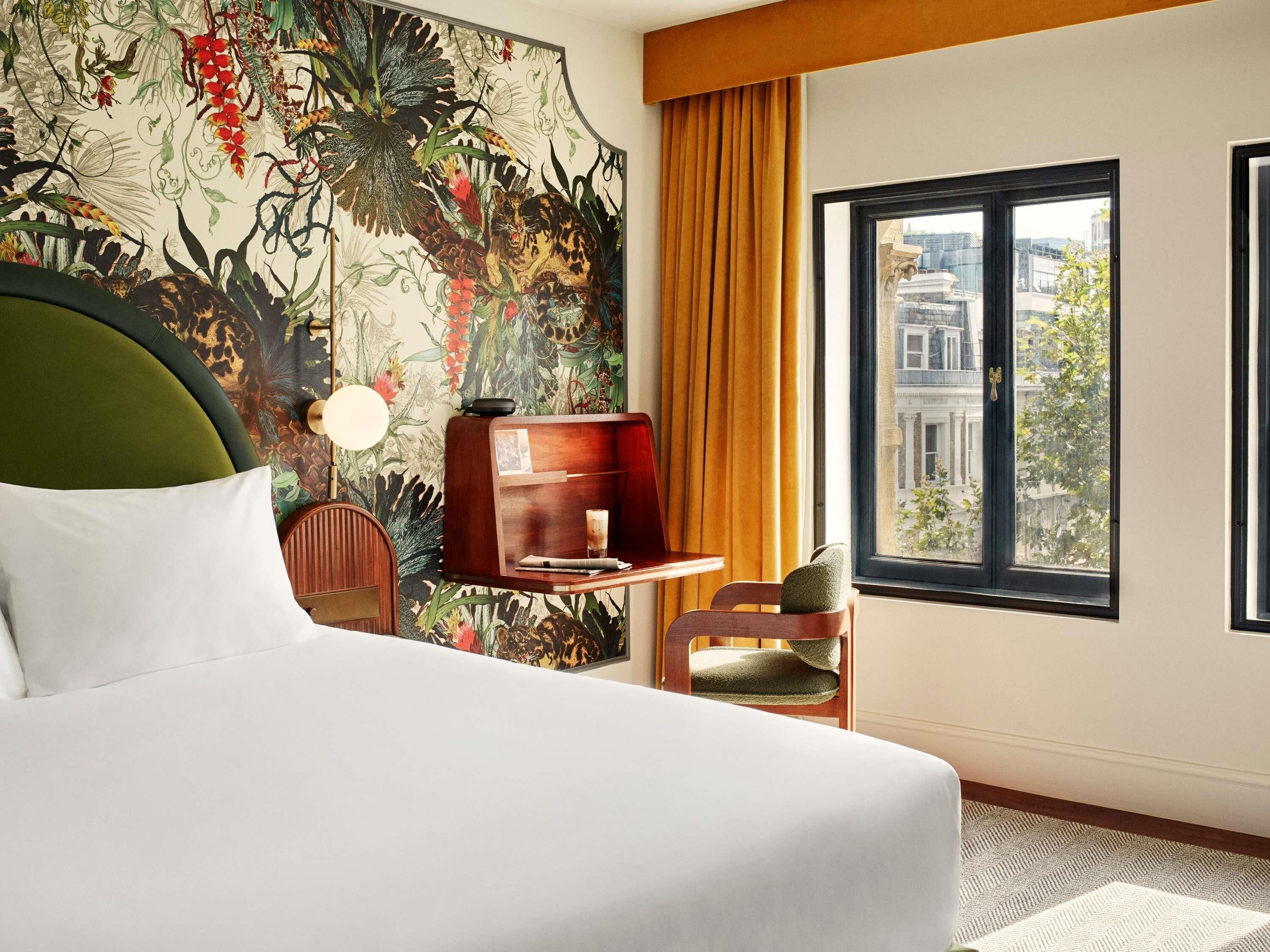 A hotel room with a bed, a desk, a chair, a large window with a city view, and a floral wallpaper.