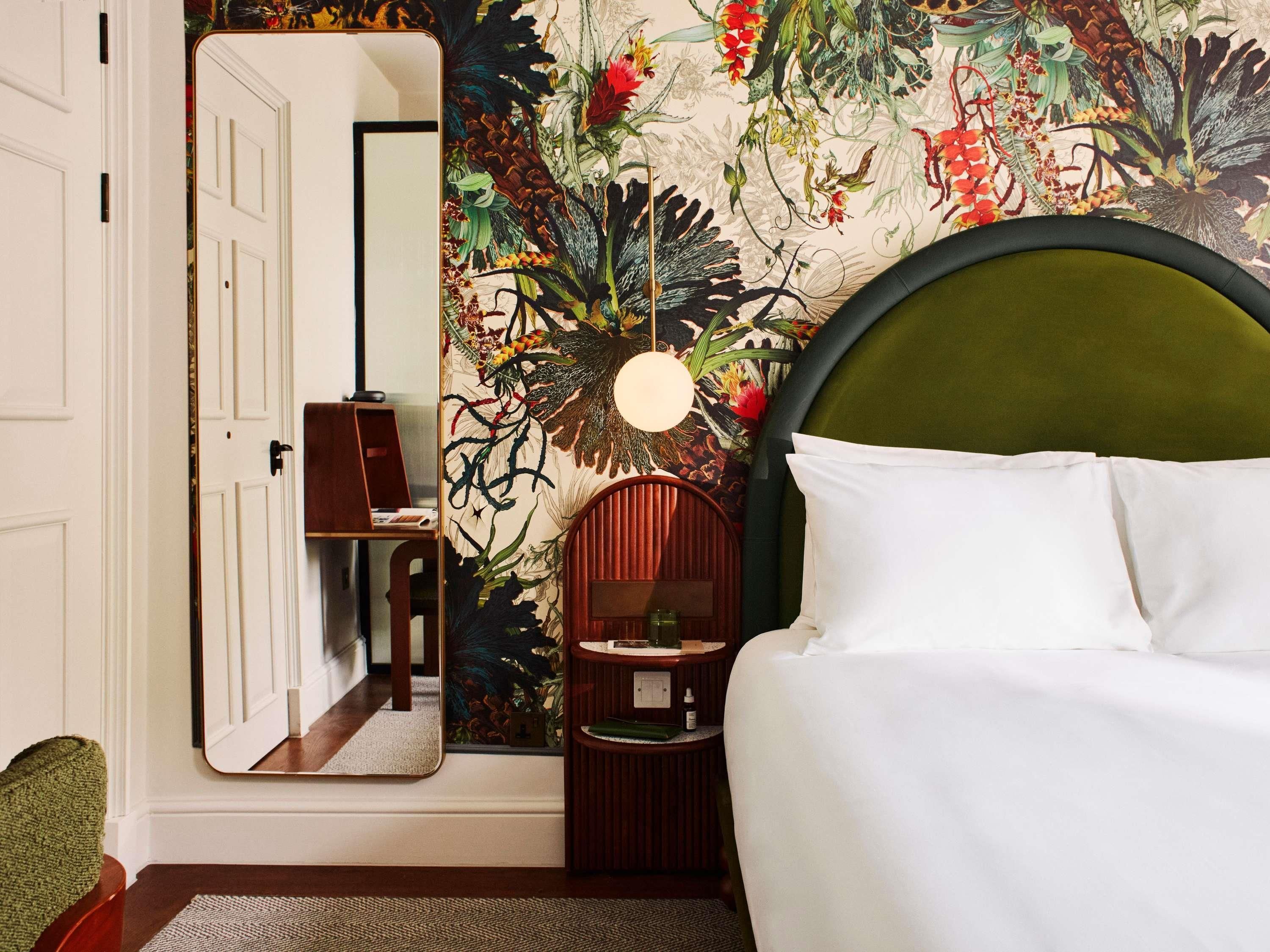 A bedroom with a bed, a bedside table, a mirror, and a wall with a floral mural.