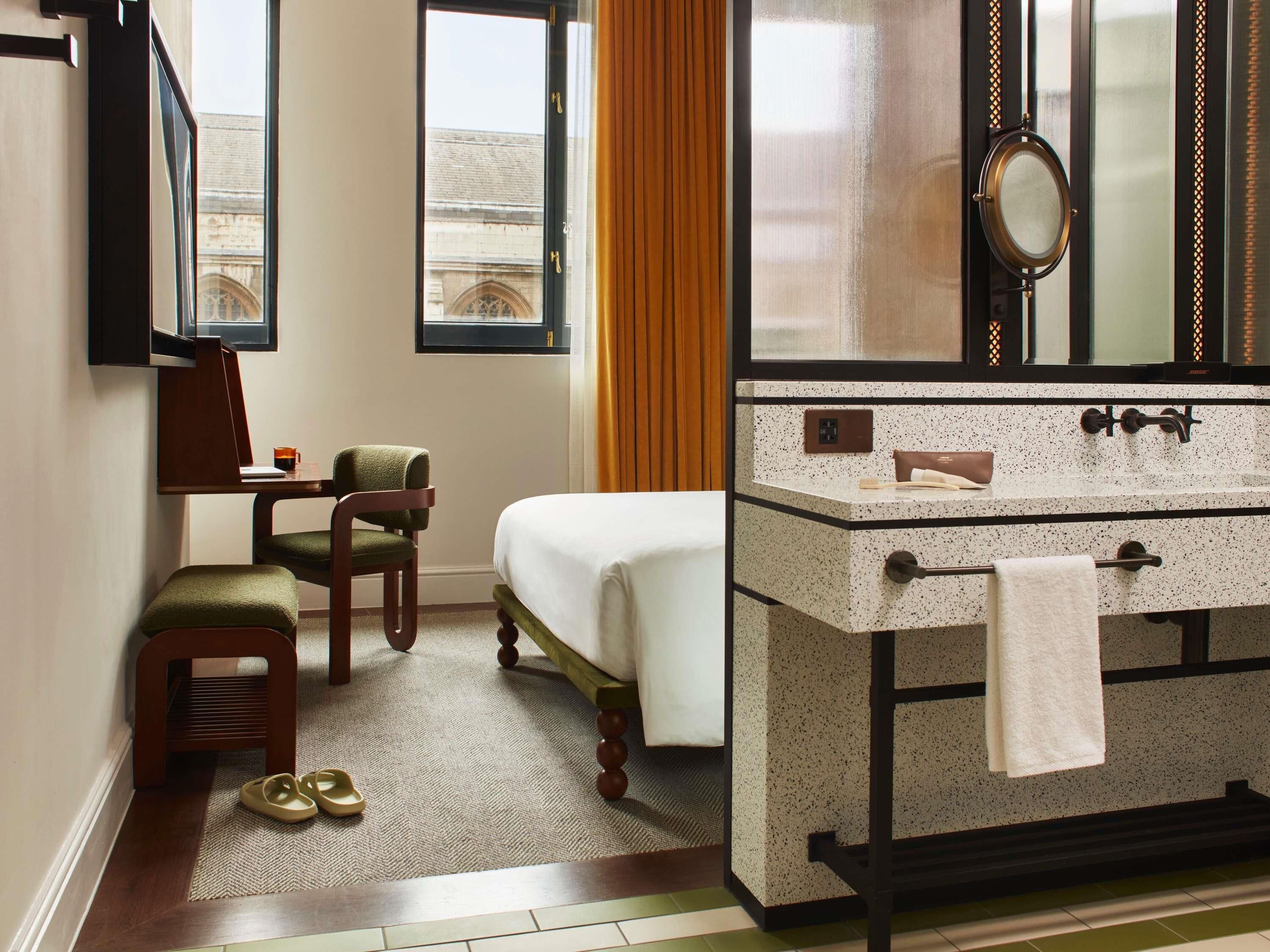 A hotel room with a bed, a desk, a chair, a bathroom with a sink and mirror, and a view of a building outside.