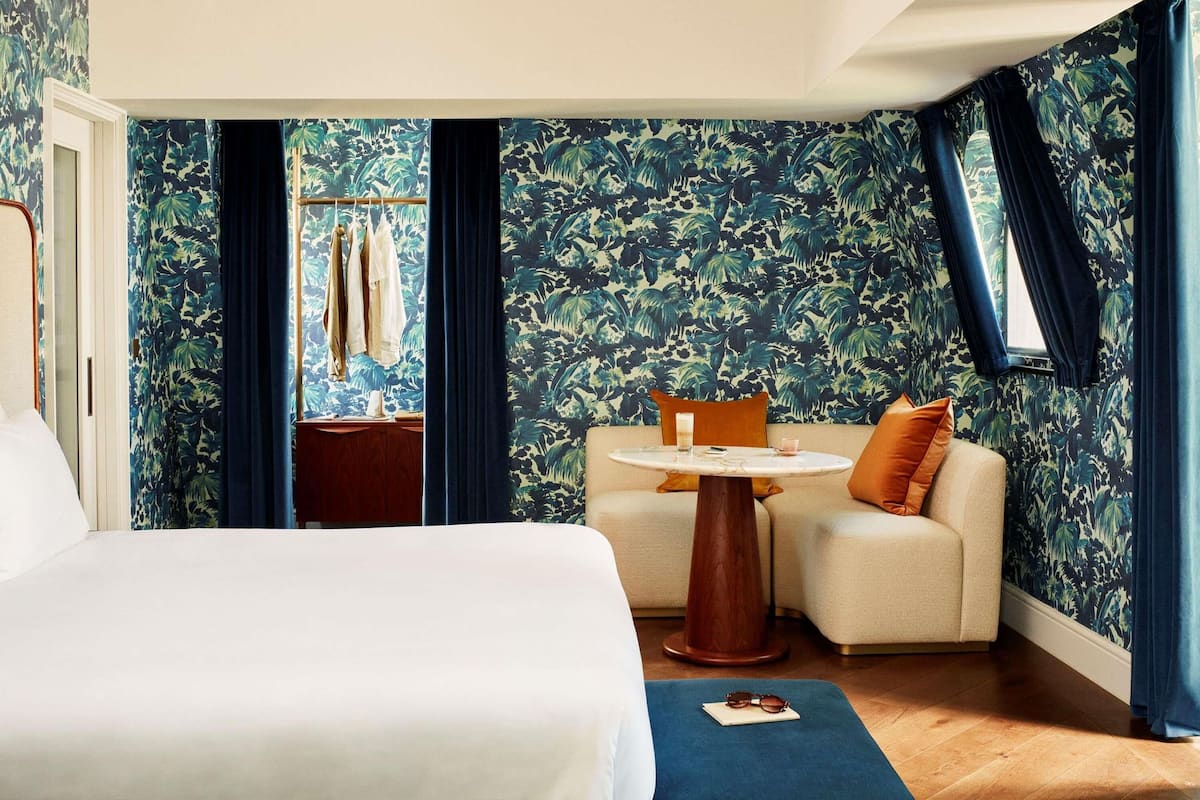 Une chambre d’hôtel avec un lit, une petite table ronde et une chaise, ainsi qu’un canapé. Il y a une fenêtre avec des rideaux, et les murs sont recouverts d’un motif de feuilles tropicales.