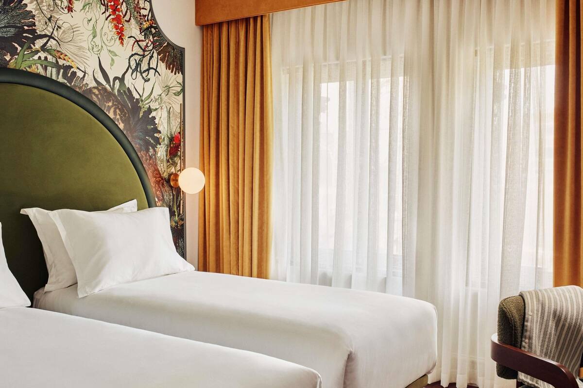 Une chambre d’hôtel avec deux lits, une grande tête de lit, une lampe fixée au mur et une fenêtre avec des rideaux légers.