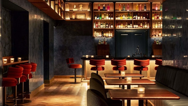 Uma área de bar com estantes de madeira, banquetas vermelhas e mesas com velas.
