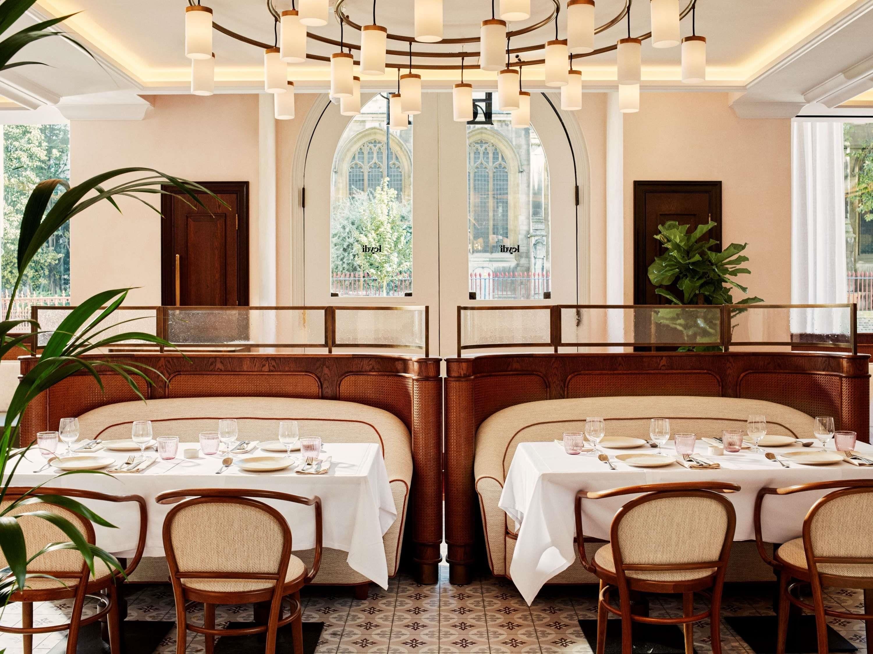 A restaurant with a high ceiling, hanging pendant lights, and a view of a church through arched windows.