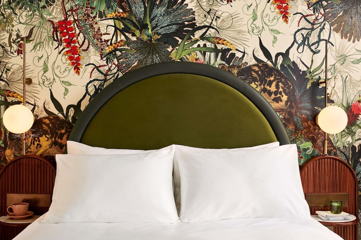 Un lit avec du linge de lit blanc, une tête de lit recouverte d’un tissu vert et un mur orné d’une fresque représentant des motifs floraux tropicaux.