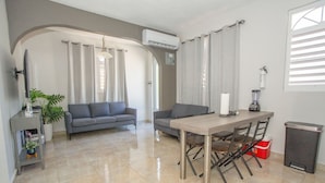 A modern living room with a sofa, a dining table with chairs, a TV, and a plant. - EcoMar Rentals Apartment 3/2 (Cabo Rojo)