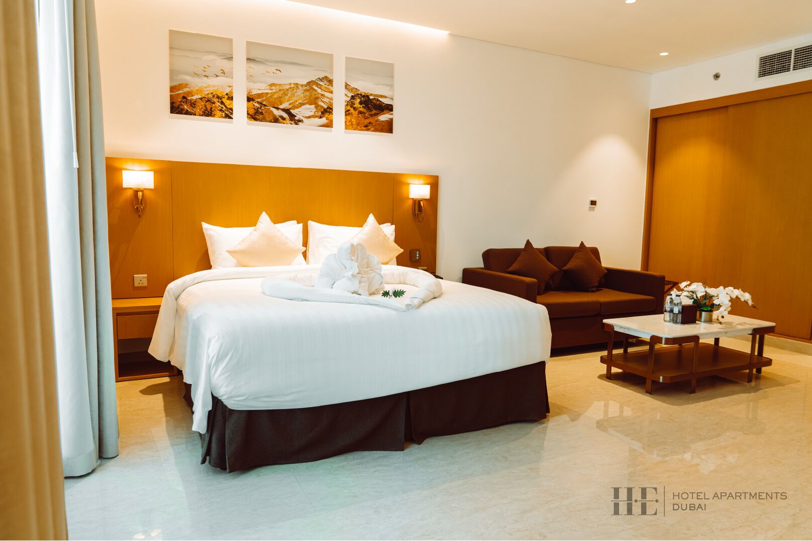 A hotel bedroom with a large bed, a sofa, a small table with flowers, and wall-mounted artwork.