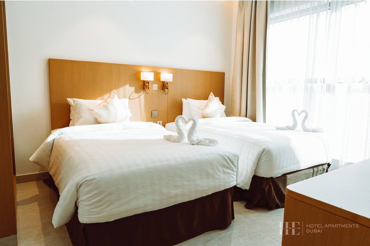 A hotel room with two beds, a wooden headboard, wall-mounted lamps, and a view of the city.