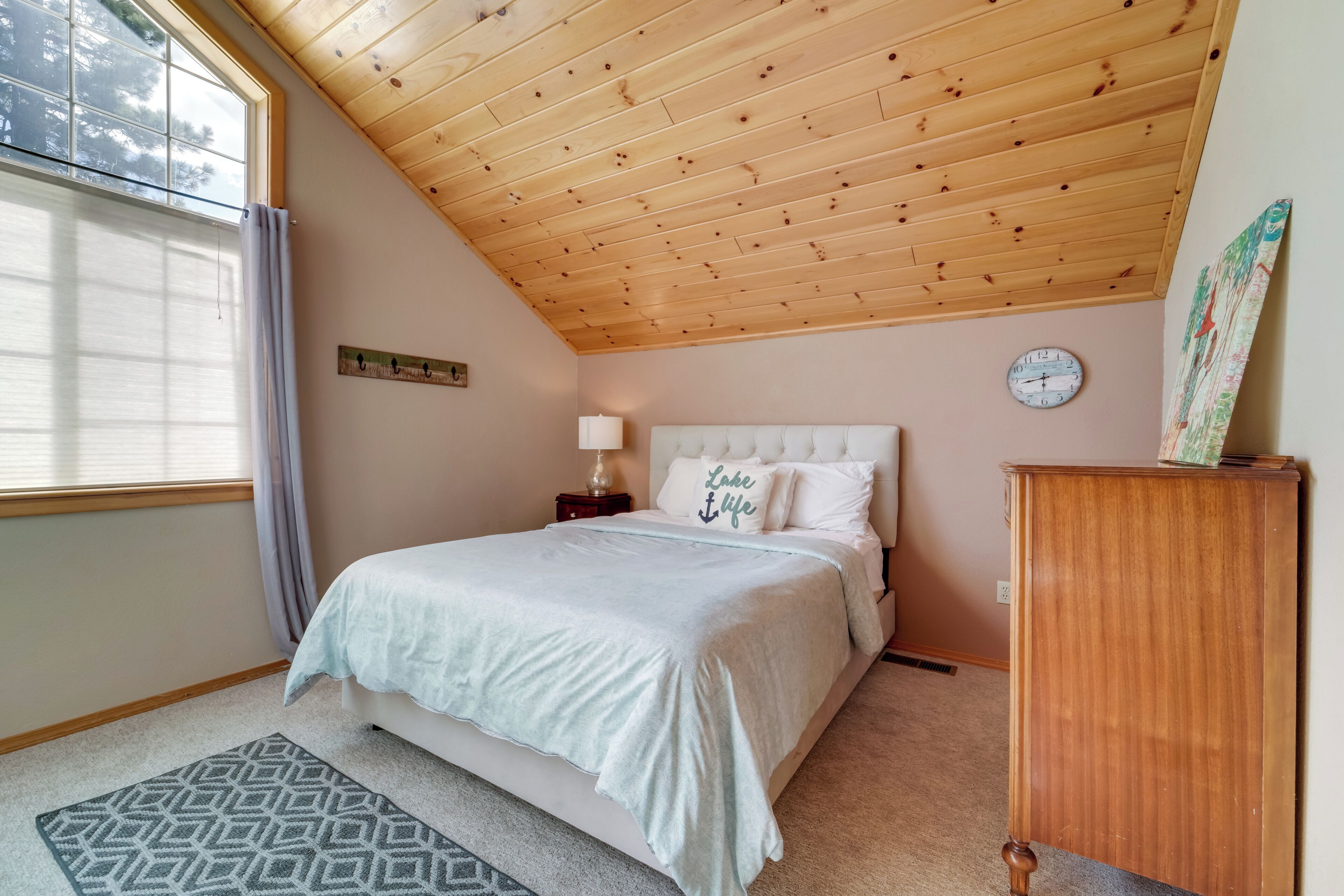A bedroom with a wooden ceiling, a bed with a white headboard, a clock on the wall, and a small wooden dresser.
