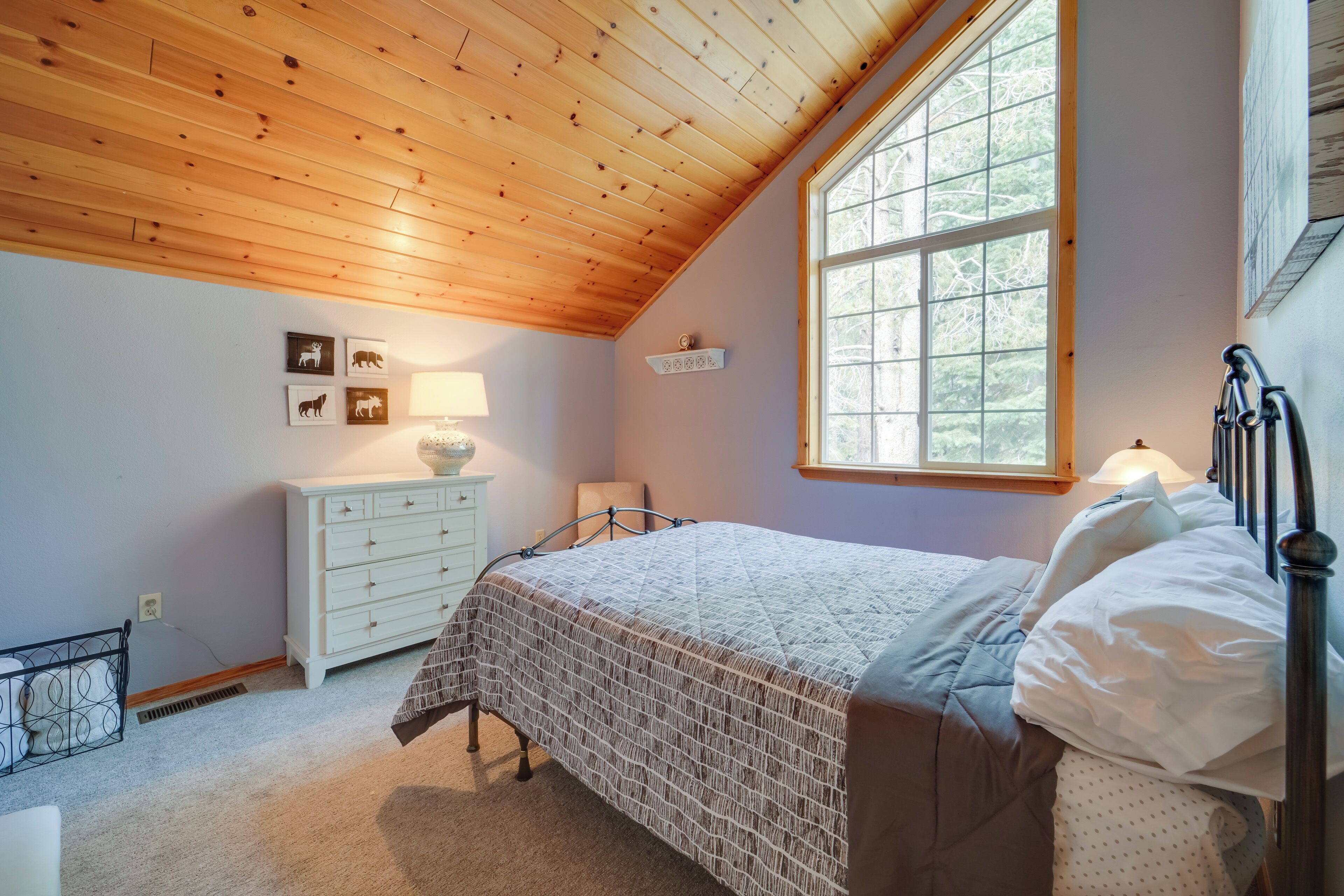 A bedroom with a wooden ceiling, a large window, a bed with a quilted bedspread, a nightstand with a lamp, and wall-mounted artwork.
