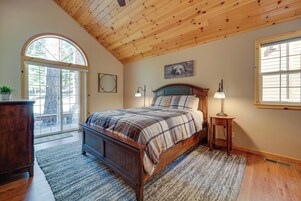 Quarto com teto de madeira, uma cama grande com roupa de cama xadrez, mesa de cabeceira com abajur e janela com persianas.