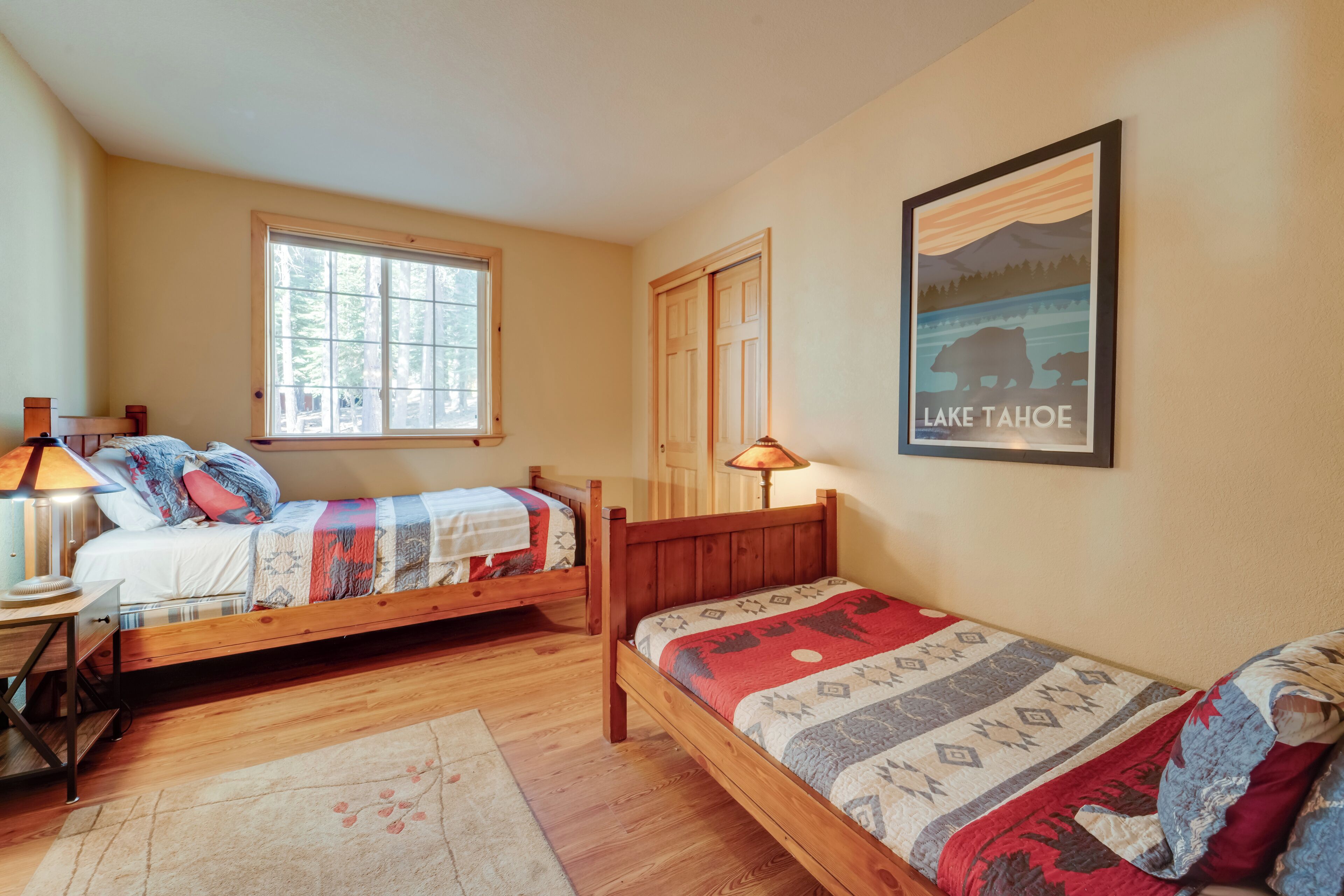 A bedroom with two wooden beds, a window, a lamp, a rug, and a poster of Lake Tahoe.