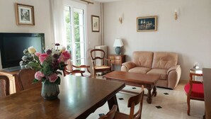 Een woonkamer met een bank, een eettafel, stoelen, een televisie en een vaas met bloemen.