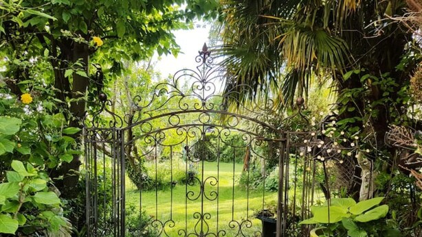Een ijzeren hekwerk met ingewikkelde decoraties, dat uitkomt op een weelderige tuin met diverse planten en een verzorgd gazon.