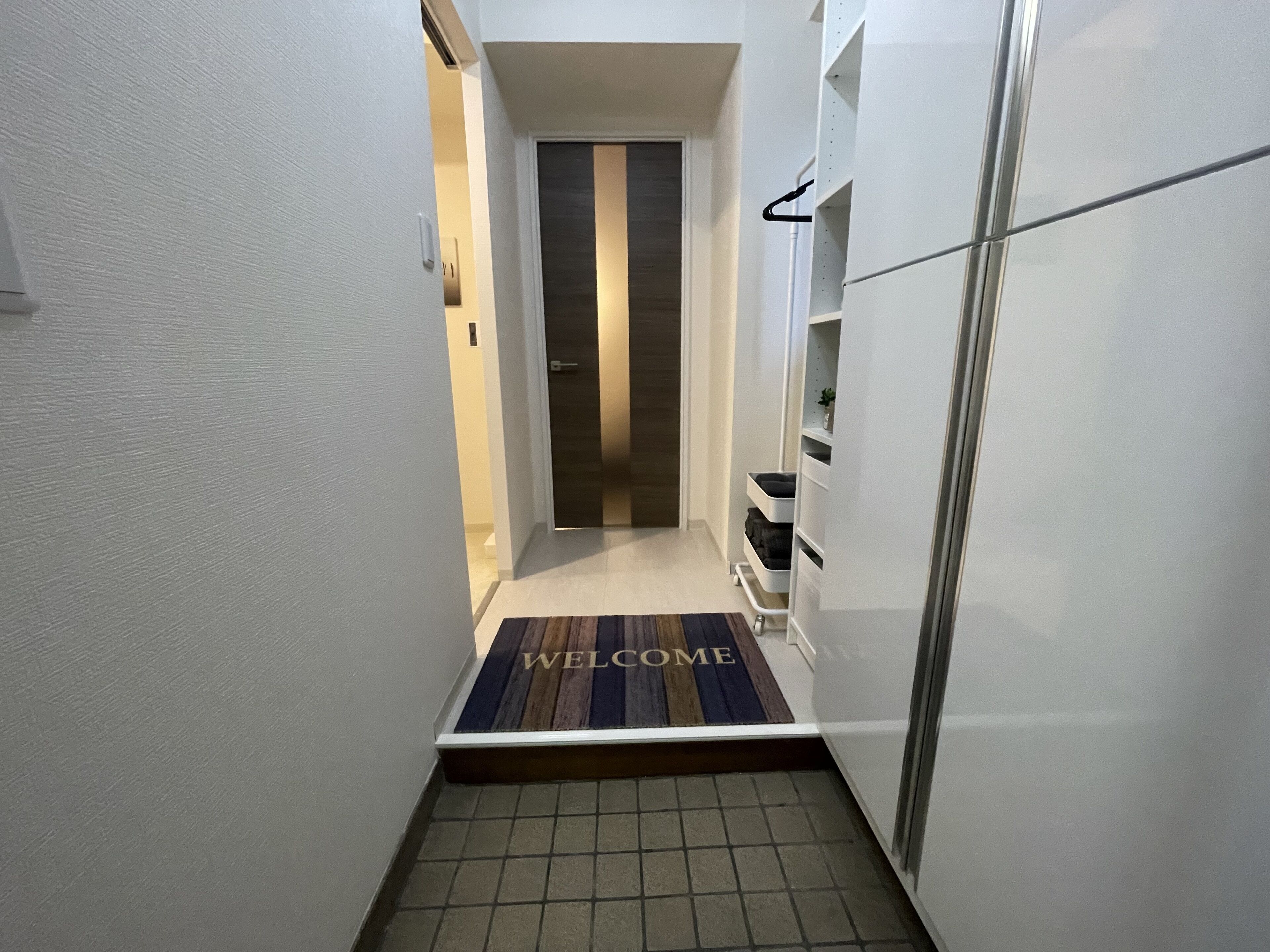 A narrow hallway with a welcome mat, a door at the end, and a white cabinet on the right.