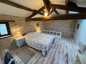 Un dormitorio con pared de piedra, vigas de madera, una cama con colcha estampada, lámparas de noche, un radiador y una ventana con vistas.