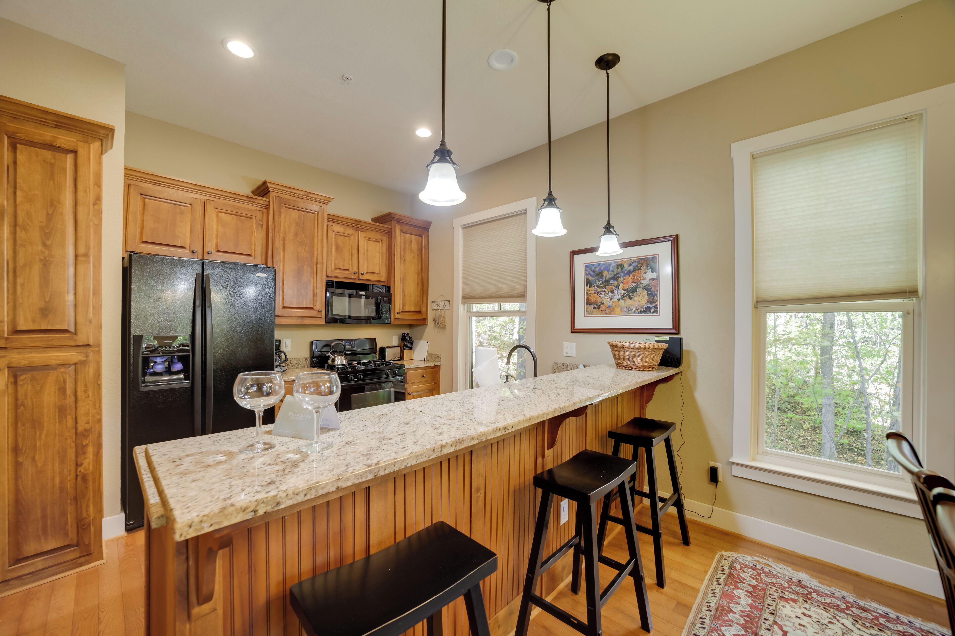 A kitchen with wooden cabinets, a black refrigerator, a granite countertop, pendant lights, and a window with a view of trees.