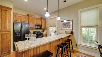 A kitchen with wooden cabinets, a black refrigerator, a granite countertop, pendant lights, and a window with a view of trees.
