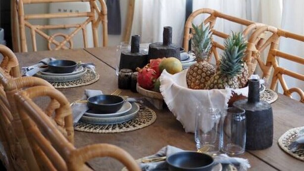 Ein Essbereich mit einem Holztisch, gedeckt für zwei Personen, mit gewebten Platzsets, einer Tischdekoration mit Ananas und hängenden Lampen aus Netz.