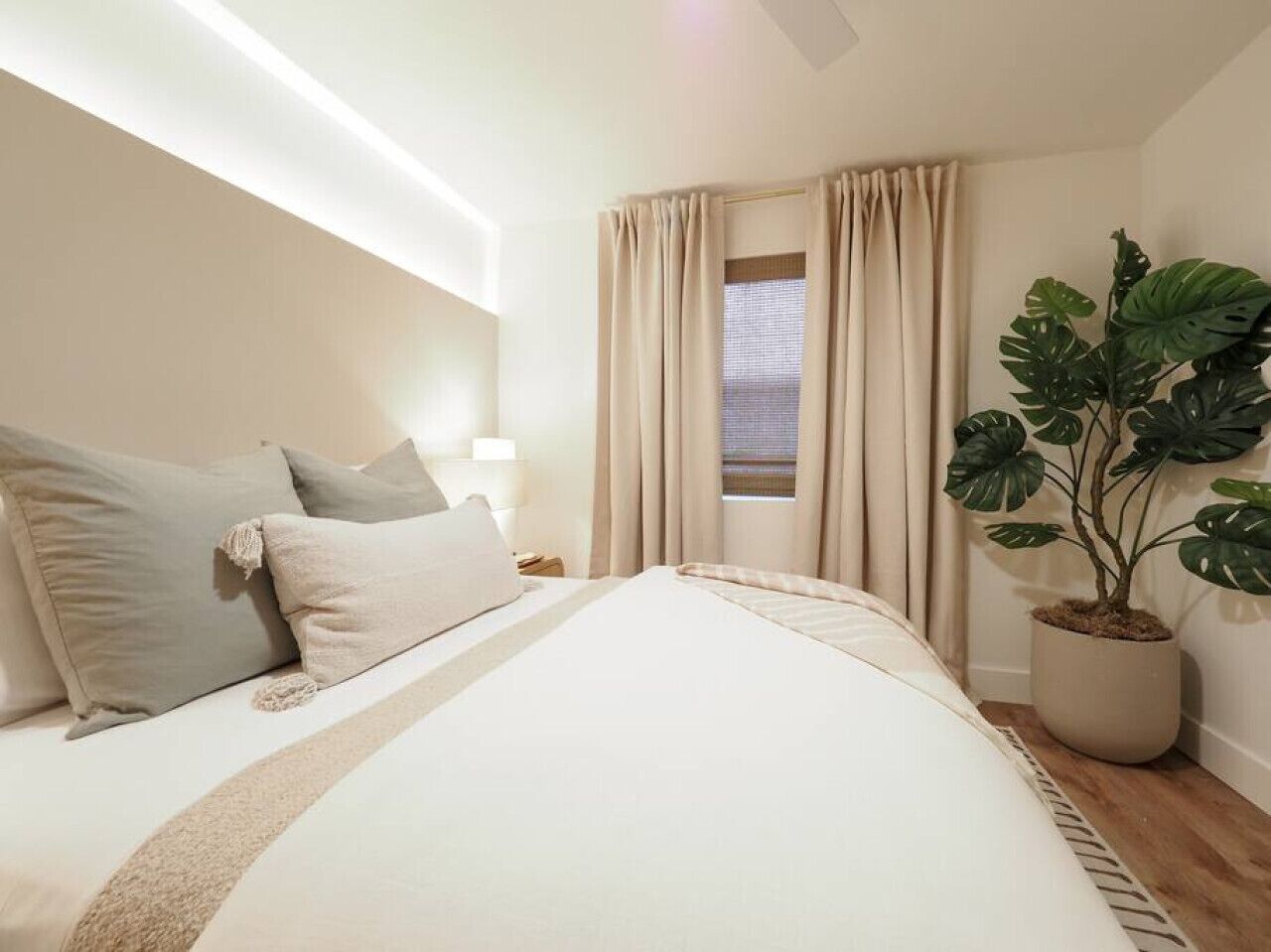 Quarto com uma cama grande, cortinas bege, uma planta em vaso e uma mesa de cabeceira com abajur.