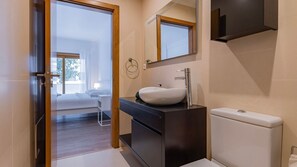 Ein modernes Badezimmer mit einem weißen Waschbecken, einer dunklen Kommode und einer Toilette. Ein Spiegel spiegelt einen Teil des Raumes wider, und eine Tür führt in ein Schlafzimmer mit Bett und Fenster.