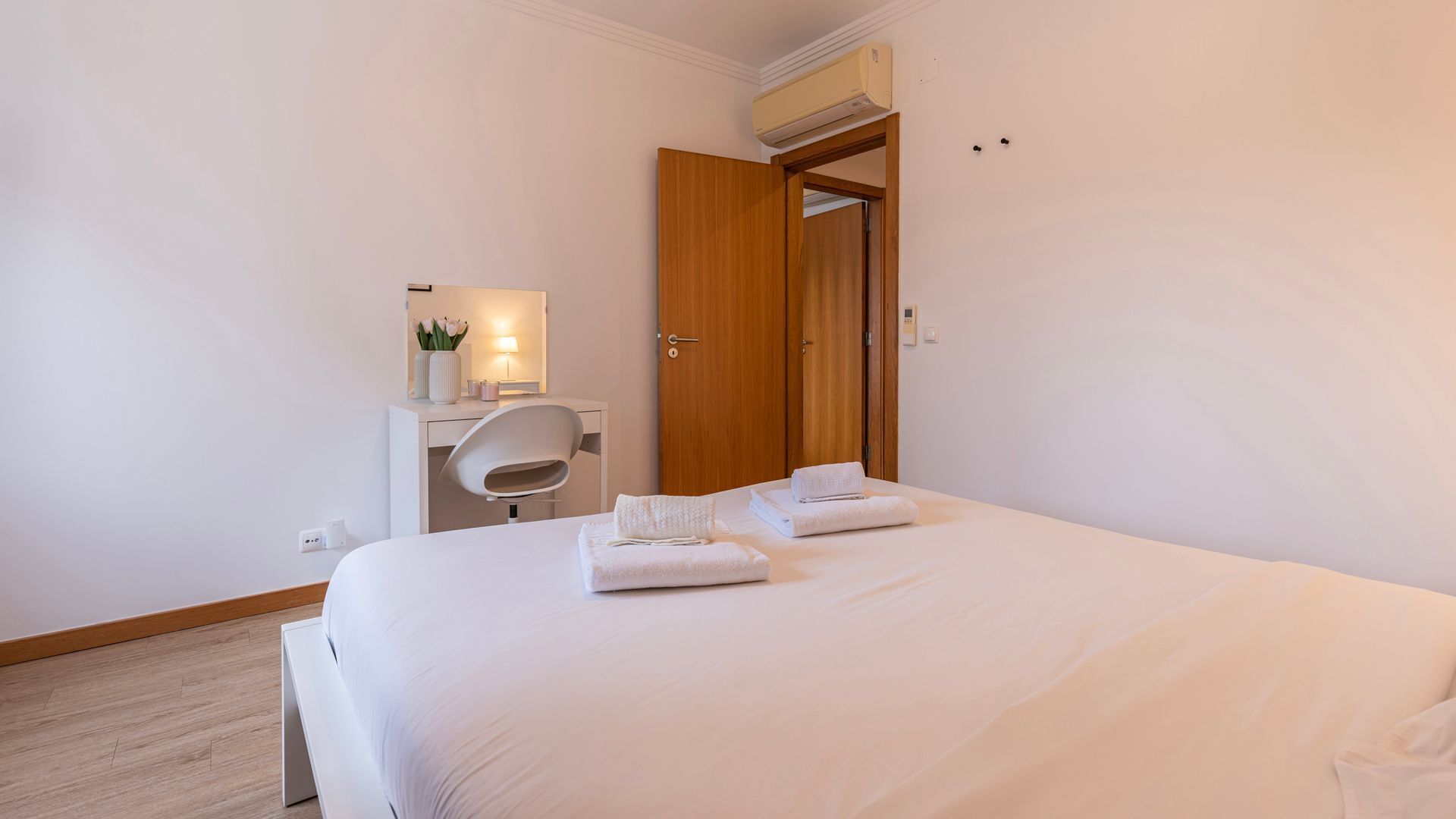 A hotel room with a large bed, a desk with a chair, a wooden wardrobe, and a small table with a lamp and flowers.
