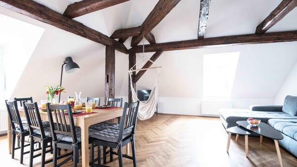 A dining area with a wooden table and chairs, a hanging hammock, and a sofa in a loft-style room with exposed wooden beams.