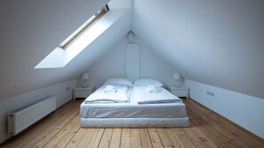 A bedroom with a sloped ceiling, a skylight, a bed with neatly folded towels, a bedside table with a lamp, and a radiator.