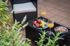 Una silla de mimbre con un cojín, una mesa con frutas, un vaso de jugo de naranja y un croissant.