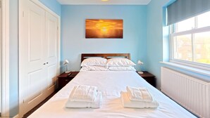 A neatly made bed with white linens and towels, a wooden headboard, two bedside lamps, and a painting of a sunset on the wall. - Stunning Coastal Cottage - Sleeping 8 in 4 Bedrooms - Pool and Beach Access Included. (Filey)