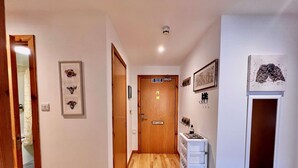 Un pasillo con suelo de madera, una pared blanca con obras de arte enmarcadas, una puerta con señal de salida de incendios y un armario blanco con ganchos.