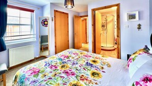 Un dormitorio con cubrecama floral, una ventana con cortinas, un radiador, una puerta de madera y un baño con ducha.