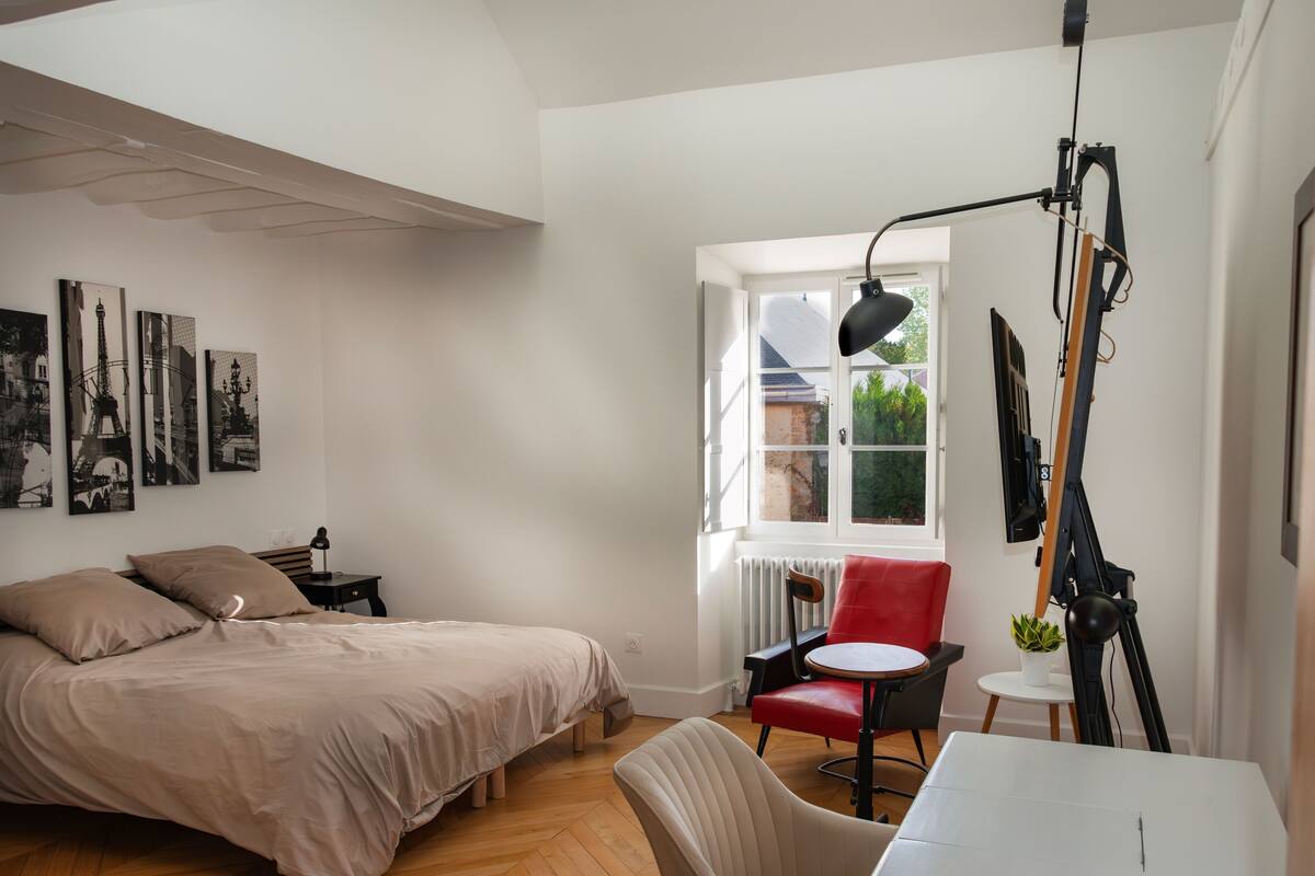 Een slaapkamer met een bed, een stoel, een tafel en een raam met uitzicht op groen.