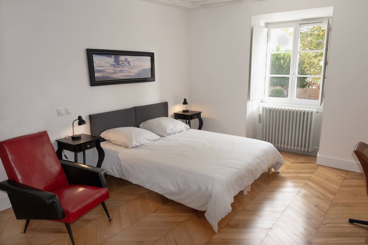 Een slaapkamer met een bed, een rode stoel, een nachtkastje, een foto aan de muur en een raam met uitzicht op groen.