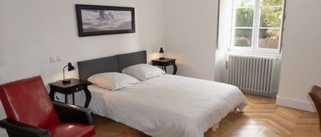 Un dormitorio con cama, una silla roja, una mesita de noche, un cuadro en la pared y una ventana con vistas a zonas verdes.