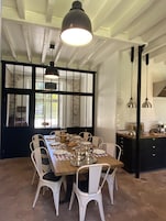Uma sala de jantar com uma mesa de madeira posta para seis pessoas, cadeiras brancas e uma luminária pendente preta acima.