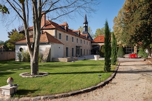 Um edifício histórico com torre do relógio, rodeado por jardins bem cuidados e uma entrada com calçada de cascalho.