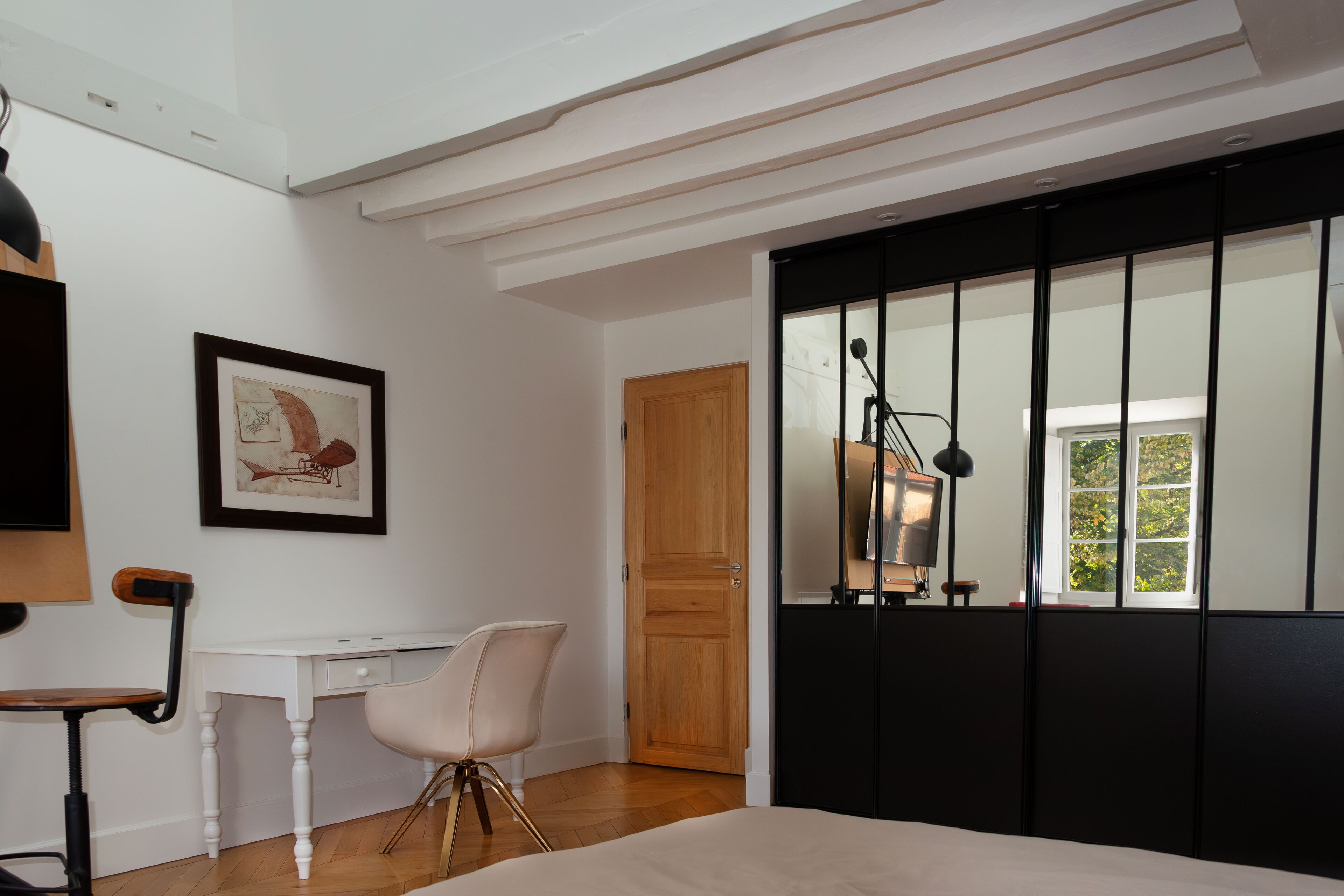A bedroom with a bed, a desk, a chair, a wooden door, and a window with blinds.