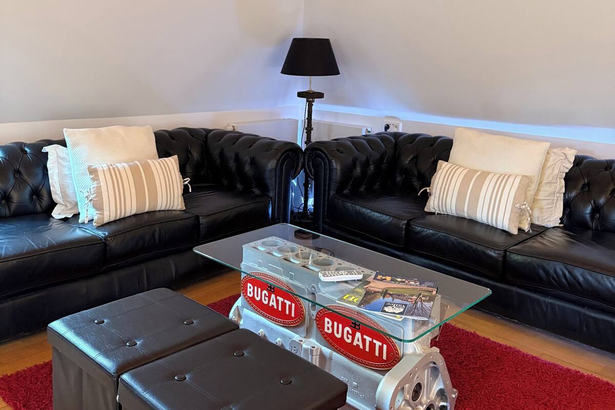 Een moderne woonkamer met een glazen salontafel waarop het Bugatti logo staat, twee zwarte leren banken met witte en gestreepte kussens, een rood vloerkleed en een zwarte vloerlamp.