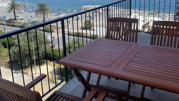 Un balcon avec une table et des chaises en bois, donnant sur une plage parsemée de parasols et de palmiers.