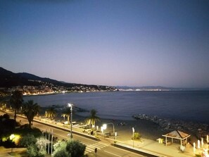A coastal cityscape at dusk with a well-lit promenade, a beach, and a mountain in the background.