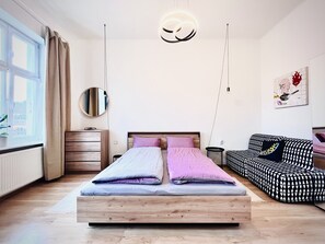 A modern bedroom with a wooden bed, a round mirror, a black and white patterned sofa, and a view of the outdoors through a window. - Sunny City View - Modern living
 (Karlovarský kraj)