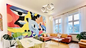 Ein modernes Wohnzimmer mit einem farbenfrohen abstrakten Wandbild, einem Glastisch, einer Couch und Blick auf die Stadt durch große Fenster.