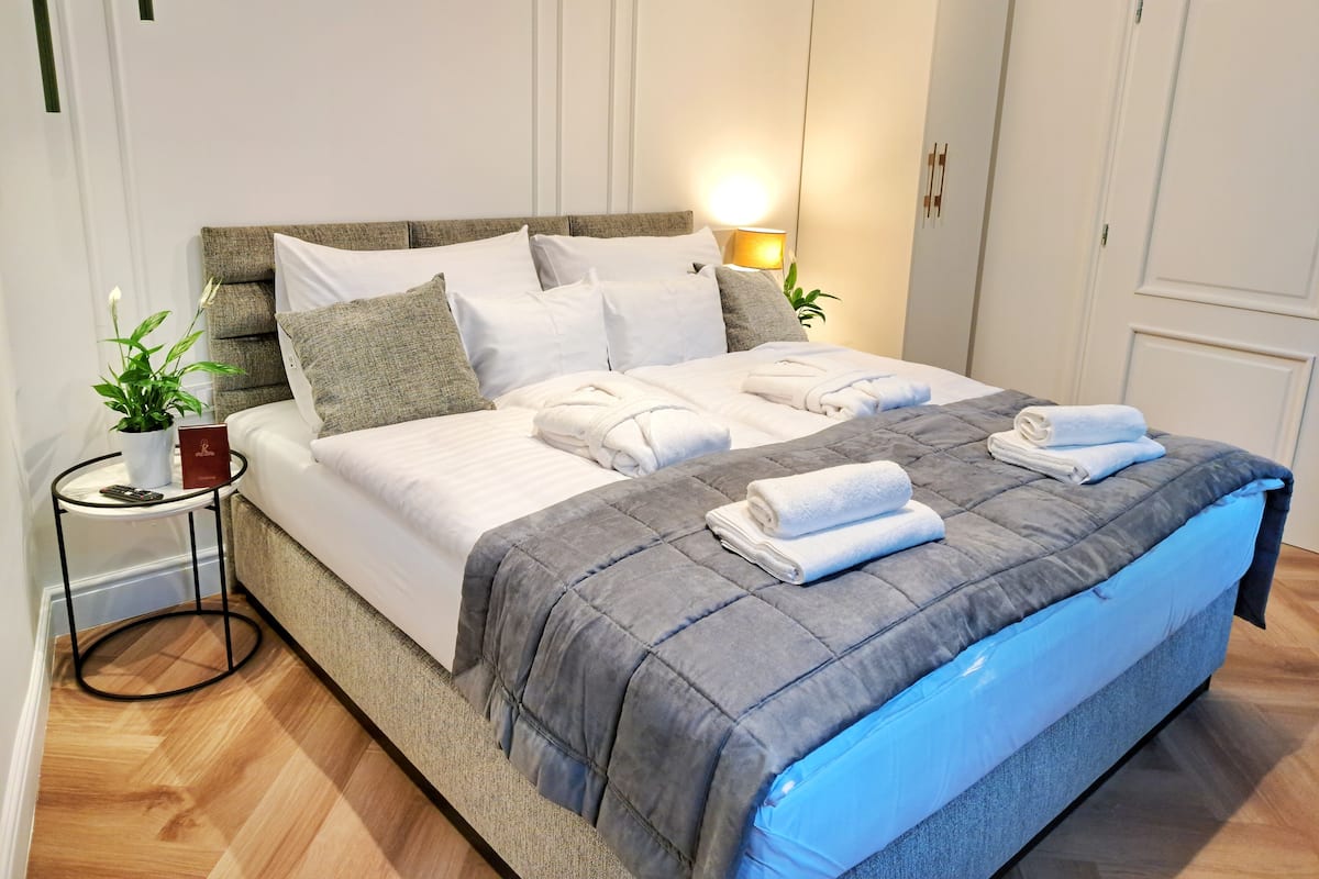A neatly made bed with white linens and a grey quilt, a bedside table with a plant and a book, and a wall-mounted lamp.