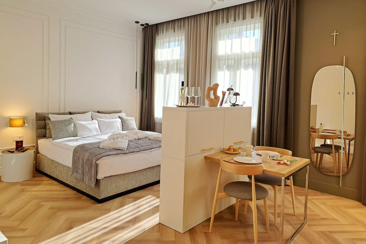 A modern hotel room with a bed, bedside table, lamp, and a breakfast setup with a table, chairs, and a mirror.